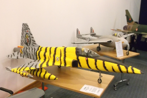 Verschiedene Flugzeugmodelle sind auf Tischen mit Etiketten angeordnet, ein Stuhl steht links und eine Wand ist im Hintergrund zu sehen.