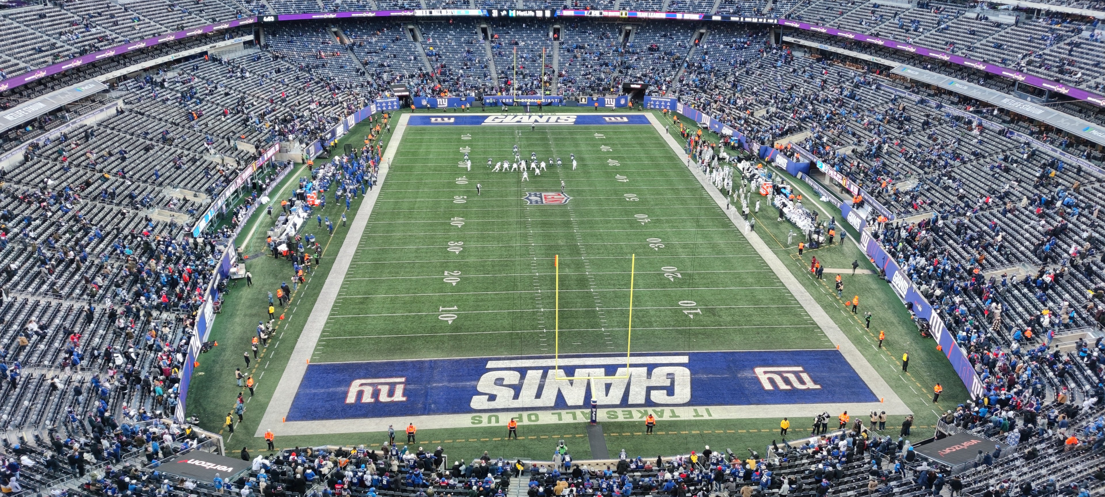 Ein überfülltes Fußballstadion mit Spielern auf dem Feld, Torpfosten und Texttafeln, aus einer erhöhten Perspektive betrachtet.