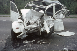 Ein stark demoliertes Auto steht am Straßenrand, mit Bäumen, einem Gebäude und einem weiteren Fahrzeug im Hintergrund.