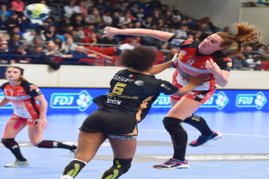 Eine Gruppe von Frauen, die auf einem Platz Handball spielen, während Zuschauer von Stühlen, Geländern und einer Wand im Hintergrund zusehen.