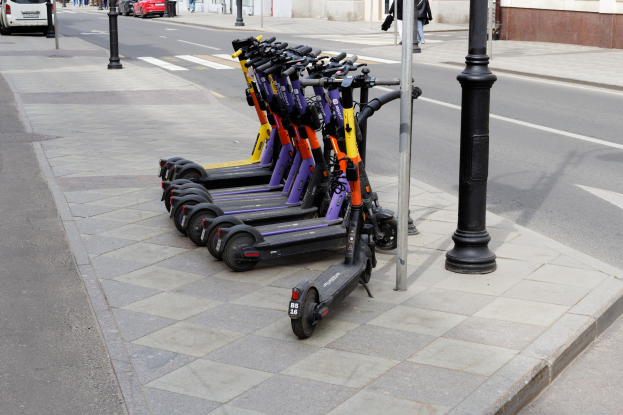 Eine Reihe von Elektro-Scootern, die an der Straßenseite geparkt sind, mit fahrenden Fahrzeugen, Menschen auf dem Gehweg und Gebäuden im Hintergrund.