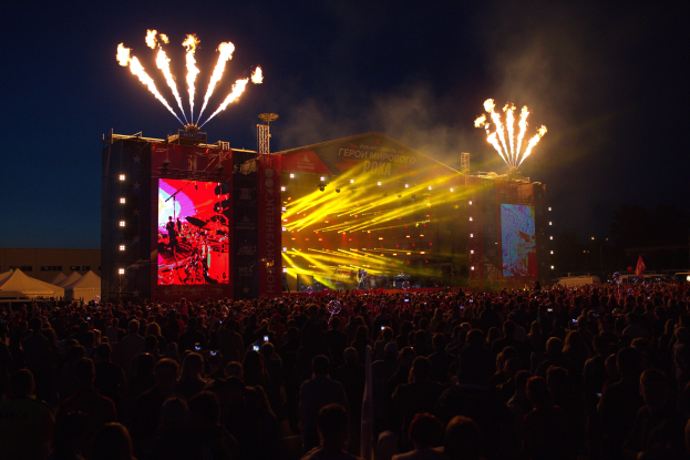 Eine große Menschenmenge auf einem Musikfestival schaut Performern auf einer hell erleuchteten Bühne zu, mit Zelten links und Feuerwerk am dunklen Himmel dahinter.
