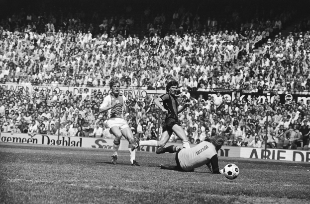 Schwarzes Foto von Männern, die auf einem Feld Fussball spielen, mit Zuschauern im Hintergrund und einer "Bundesliga 1978-79 Ajax 1-1 Hannover 96"-Fahne.