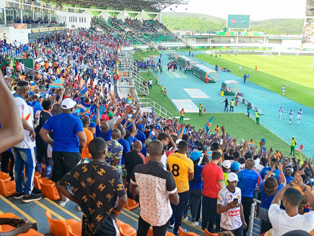 Eine große Menschenmenge versammelt sich vor einem eingezäunten Fußballfeld, wobei einige sitzen und andere stehen, unter einem klaren blauen Himmel mit Hügeln im Hintergrund, und warten auf ein Spiel.