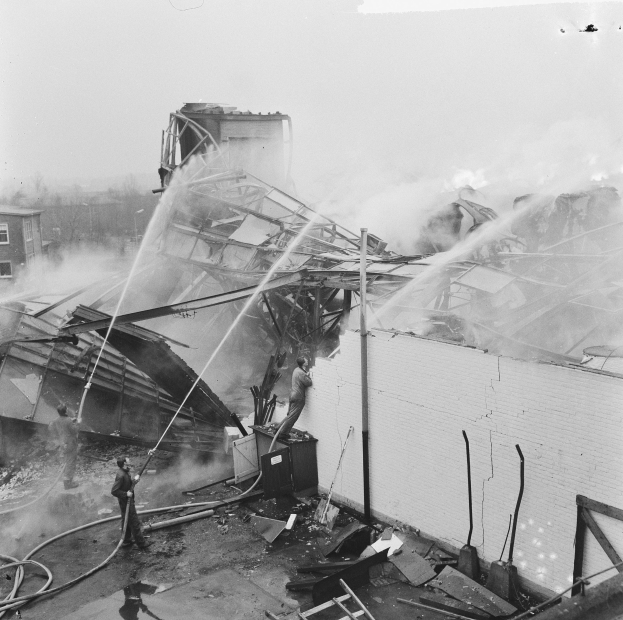 Schwarz-weiß-Foto von Feuerwehrmännern auf einem Dach, die ein Feuer löschen, während Zuschauer unten Ausrüstung halten.