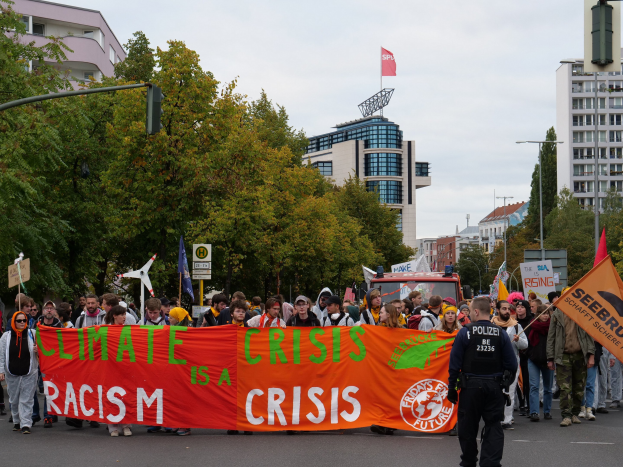 Eine Gruppe von Menschen marschiert mit einem "Klima-Krise ist eine Krise"-Schild die Straße entlang, mit Bäumen, Straßenschildern, einem geparkten Fahrzeug, Gebäuden und einem klaren blauen Himmel im Hintergrund.