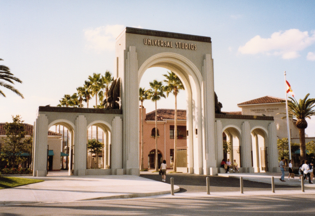 Eingang von Universal Studios Hollywood mit einem Bogen, Statue, Menschen, Bäumen, Gebäuden und einem Fahnenmast.