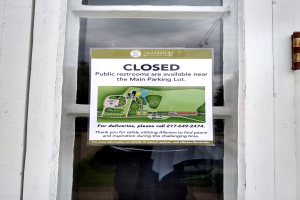 Glasstür mit einem Plakat, auf dem "Geschlossene öffentliche Toiletten in der Nähe des Hauptparkplatzes" steht und ein unklares Objekt auf der rechten Seite.