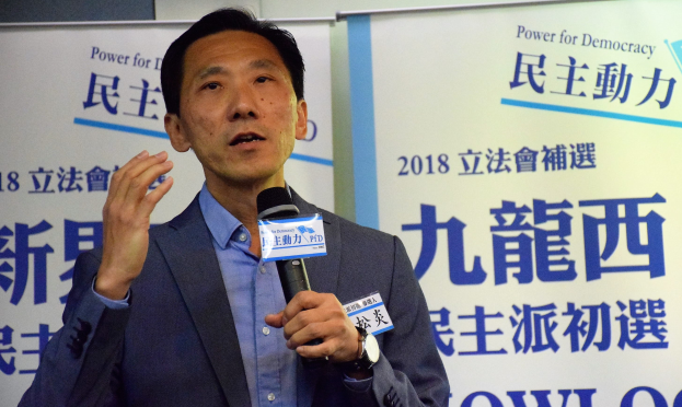 Ein Mann im Anzug spricht in ein Mikrofon mit Plakaten und einer Wand im Hintergrund und trägt eine Uhr.