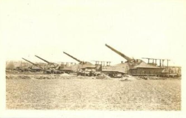 Ein Schwarz-Weiß-Foto einer Gruppe deutscher schwerer Artillerie in einem Feld, umgeben von Bäumen unter einem klaren Himmel, mit einem Rahmen versehen.