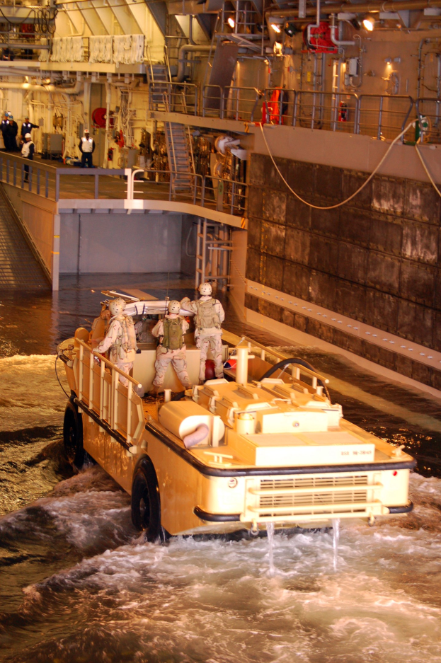 Amphibisches Fahrzeug mit Helmträgern durch Wasser neben einer Wand mit Menschen, Geländern, Lampen und Rohren im Hintergrund.