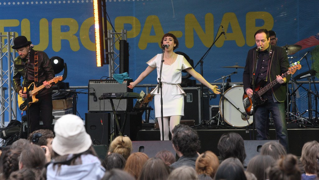 Vier Personen treten auf einer Bühne auf: eine Frau in einem weißen Kleid singt in der Mitte, während zwei Männer links und rechts von ihr Gitarre spielen; ein Publikum ist unten sichtbar, mit Lautsprechern und einer Wand im Hintergrund.