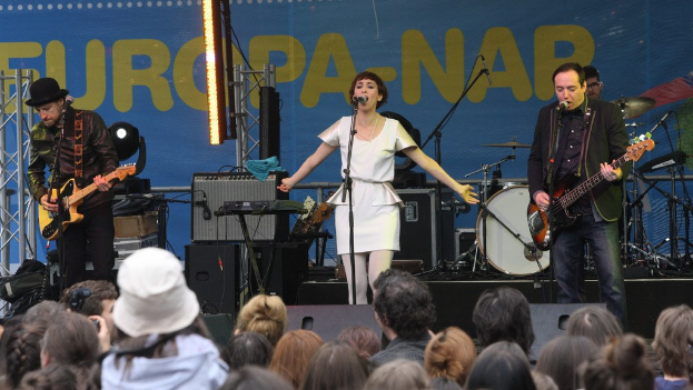 Vier Personen treten auf einer Bühne auf: eine Frau in einem weißen Kleid singt in der Mitte, während zwei Männer links und rechts von ihr Gitarre spielen; ein Publikum ist unten sichtbar, mit Lautsprechern und einer Wand im Hintergrund.