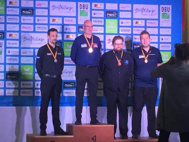 Five medal-winning men on a podium at the DDBU World Championship 2019, with a photographer capturing the moment.