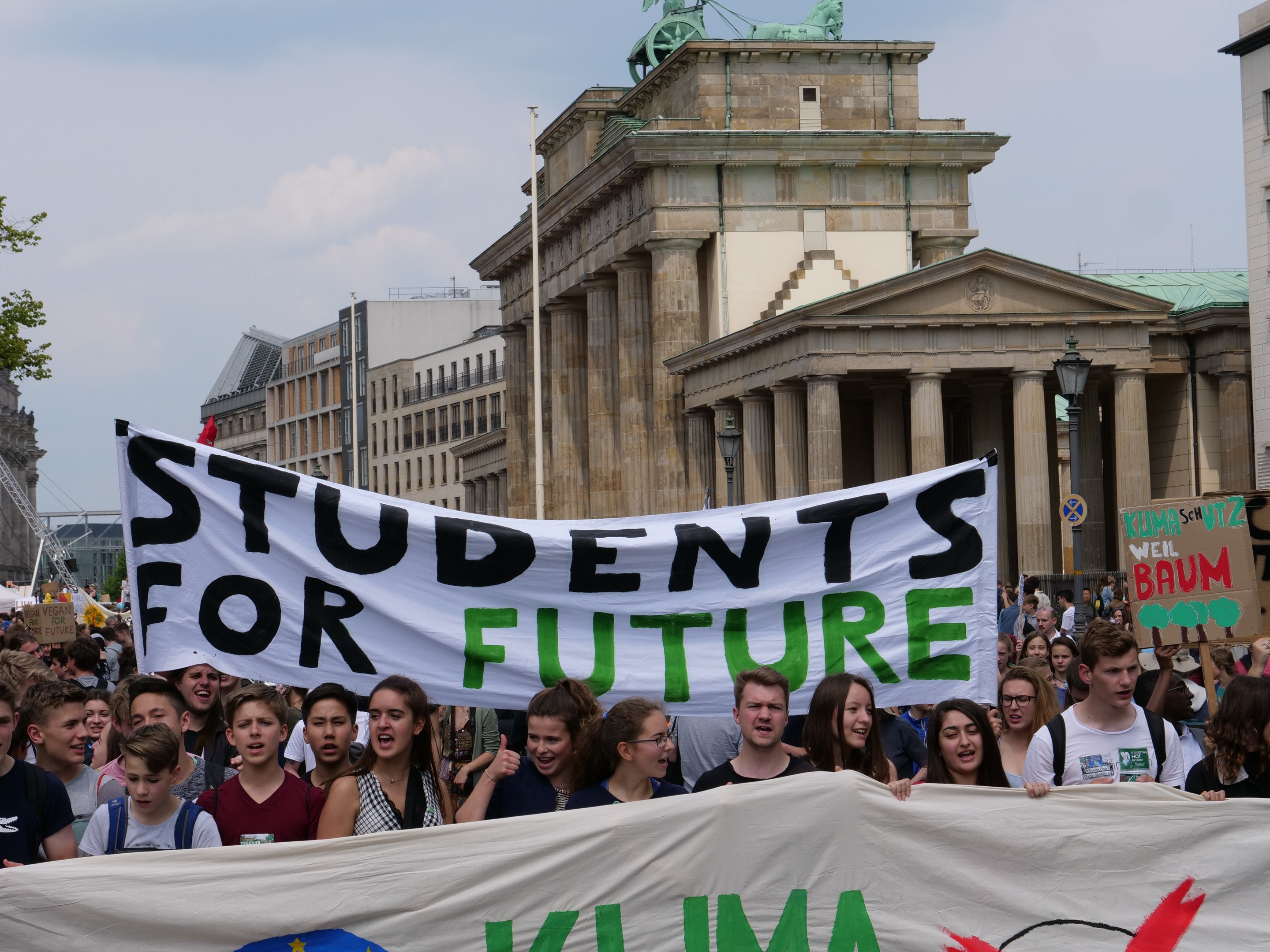 Gruppe von Schülern marschiert in Berlin mit einem bunten "Students for Future"-Schild gegen Gebäude, Bäume und Himmel.