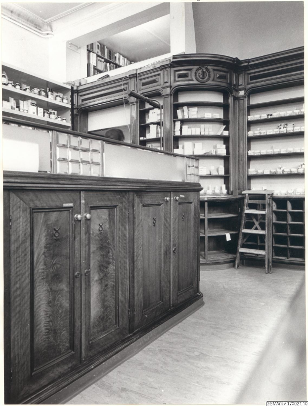 Schwarzes und wei├čes Foto eines vintage Apothekeninneren mit einer Theke, Sch├Ąben, einer Leiter und Regalen, die mit verschiedenen Gegenst├Ąnden gefüllt sind, mit Text über dem Bild.