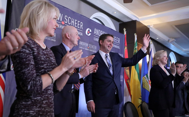 Eine Gruppe von Menschen, die vor Flaggen stehen und jubeln, mit einem Tisch voller Gegenstände rechts daneben, Stühlen, einer Fahne und einer Wand im Hintergrund sowie Lichtern und einem Ventilator auf dem Dach.