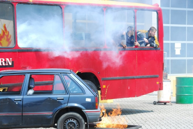 Ein roter Doppeldeckerbus mit Rauch, daneben ein geparktes Auto, drei sichtbare Passagiere und ein Gebäude mit Glasfenstern und einem Fass im Hintergrund.
