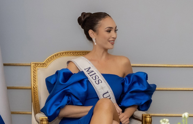 Frau in blauem Kleid mit einem "Miss Universe 2019"-Band sitzend auf einem Stuhl umgeben von Blumen mit einem Lächeln im Gesicht vor einer Wand.