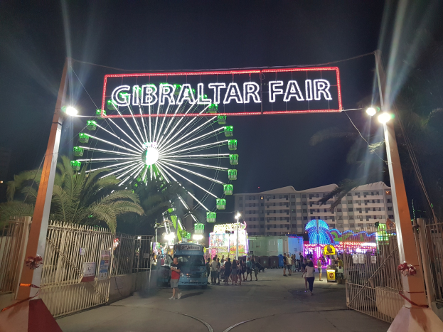 Der Eingang zur Gibraltar-Messe bei Nacht, beleuchtet von einem großen Riesenrad und einer leuchtenden Tafel, mit Menschenmassen drumherum, Bäumen und Gebäuden im Hintergrund unter einem dunklen Himmel.