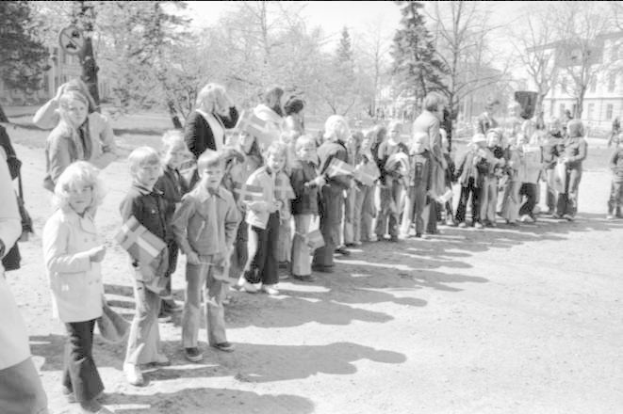 Schwarzes und weißes Bild einer Protestdemonstration auf einem Schulgelände, bei der Menschen mit Fahnen in einer Reihe auf einem Schotterweg stehen, Bäume und Gebäude im Hintergrund unter einem klaren Himmel.