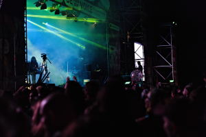 Eine volle Konzerthalle mit Performern auf einer hell erleuchteten Bühne, mit Musikinstrumenten, Mikrofonen und einem Banner im Hintergrund.