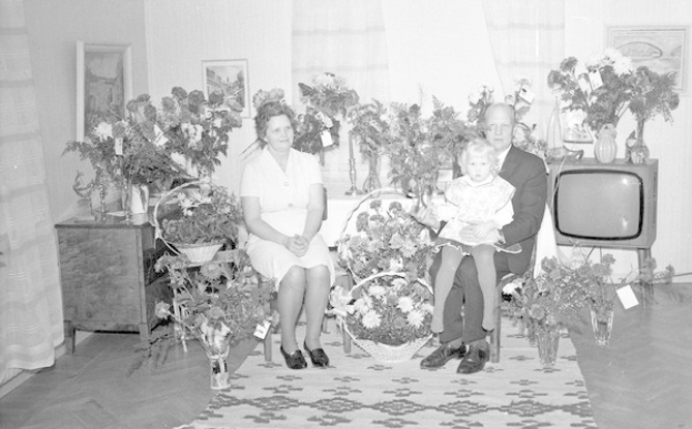 Schwarzes und weißes Foto einer Familie aus drei Personen - einem Mann, einer Frau und einem Kind - auf einem Sofa in einem Wohnzimmer mit Blumenvasen, einem Teppich, Vorhängen und Fotorahmen im Hintergrund.