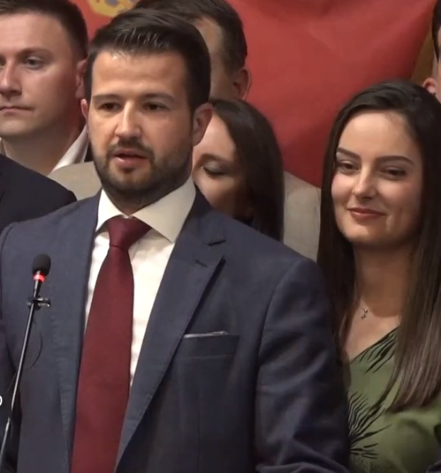 Ein Mann im Anzug und Krawatte steht an einem Rednerpult und spricht zu einer Menge mit einem Banner im Hintergrund.