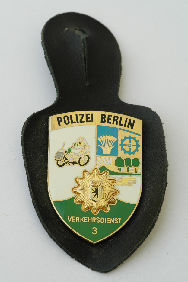Ein schildförmiges Polizeibadge mit "Polizei Berlin" in fetter schwarzer Schrift auf einem blauen Hintergrund mit einem weißen Stern und Rahmen, das auf einer Oberfläche liegt.