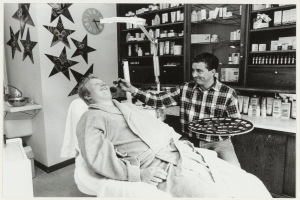 Ein Mann lächelt in einem Zahnarztstuhl, während ein anderer Mann neben ihm steht und ein Tablett hält; ein Waschbecken mit einem Wasserhahn und einer Schüssel ist links zu sehen, und Regale mit Flaschen im Hintergrund, mit Sternen und einer Uhr an der Wand dahinter.