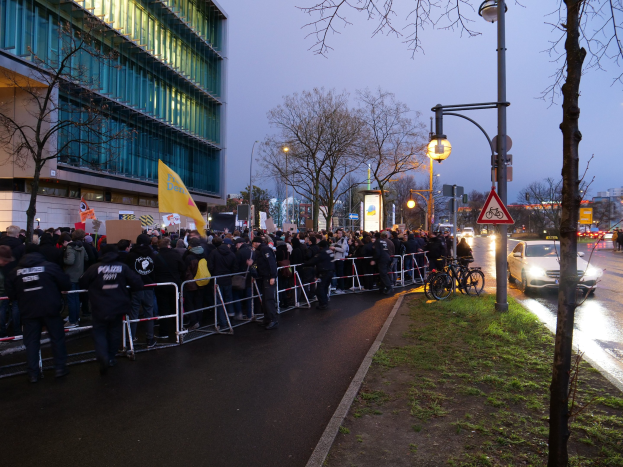 Große Gruppe von Menschen mit Schildern vor einem Gebäude während einer Demonstration in Berlin, mit Barrikaden, Fahrrädern, Laternenmasten, Schildern, Bäumen und Gras im Hintergrund.