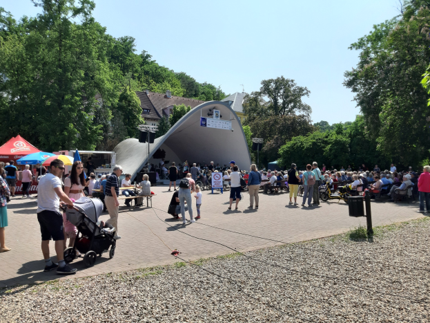 Menschenmenge vor einem Gebäude bei der Festival-Eröffnung, einige sitzen auf Bänken, andere stehen, einschließlich eines Kinderwagens im Vordergrund, umgeben von Bäumen, Gebäuden und einem klaren blauen Himmel, mit Zelten links und einem Mülleimer rechts.