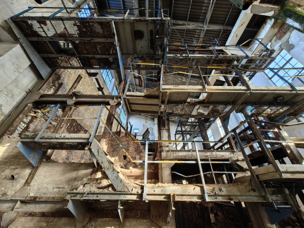 Alter Industriebau mit einem zentralen Metallgerüst, verrosteten Wänden, verfallendem Boden und umgebenden Eisenstangen, Treppen und Fenstern.