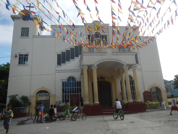 Eine Gruppe von Menschen auf Fahrrädern vor einer weißen Kirche mit architektonischen Details wie Fenstern, Säulen, Bögen, Türen und einem Kreuzsymbol, mit Pflanzen, einem Zaun, einer Fahne, Bäumen und einem bewölkten Himmel im Hintergrund und bunten Fahnen, die an einem Seil oben befestigt sind.