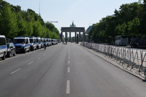 Lange Reihe von Polizeiwagen, die an einer Straße vor dem Brandenburger Tor geparkt sind, mit Fahrradfahrern und Fußgängern in der Nähe, Absperrungen, Bäume, ein prächtiges Tor mit Statuen im Hintergrund und Himmel darüber.