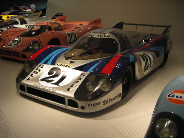 Ein Porsche 917K Rennsportwagen in einem Museum ausgestellt, umgeben von anderen Sportwagen und Menschen, mit einer Wand und einem informativen Schild im Hintergrund.