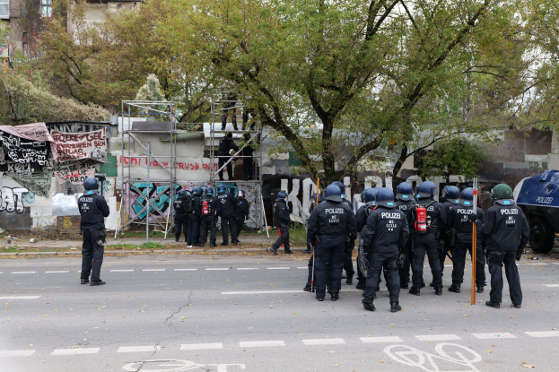 Polizisten in schwarzen Uniformen und Helmen stehen vor einem graffiti-bedeckten Gebäude, einige halten Stöcke, mit Bäumen, Gebäuden, einem blauen Himmel und einem geparkten Fahrzeug im Hintergrund.