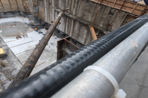 Eine Baustelle mit einem großen Metallrohr in der Mitte, umgeben von Arbeitern, Holzobjekten und verschiedenen Geräten, mit Fahrzeugen und einem Gebäude im Hintergrund.