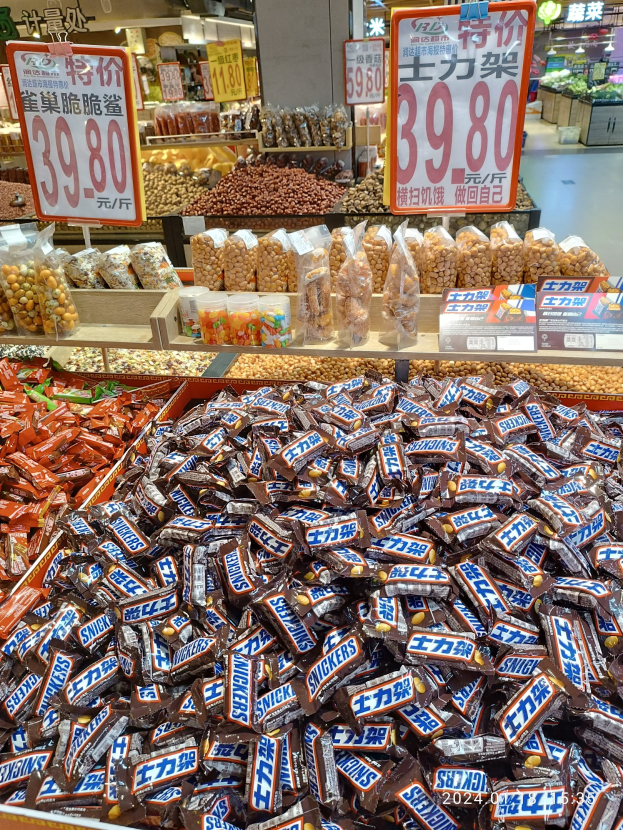 Vitrine mit verschiedenen Sorten von Süßigkeiten, einschließlich Snickers, in einem Geschäft mit Lebensmitteln, Regalen, Tafeln und Deckenlampen im Hintergrund.