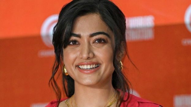 Eine Frau in einem roten Kleid mit einem eleganten Dutt, schwarzen High Heels und natürlichem Make-up lächelt in die Kamera, mit einem hellgelben Banner im Hintergrund, auf dem 'Sonam Kapoors Schönste Looks vom Cannes Film Festival' steht.