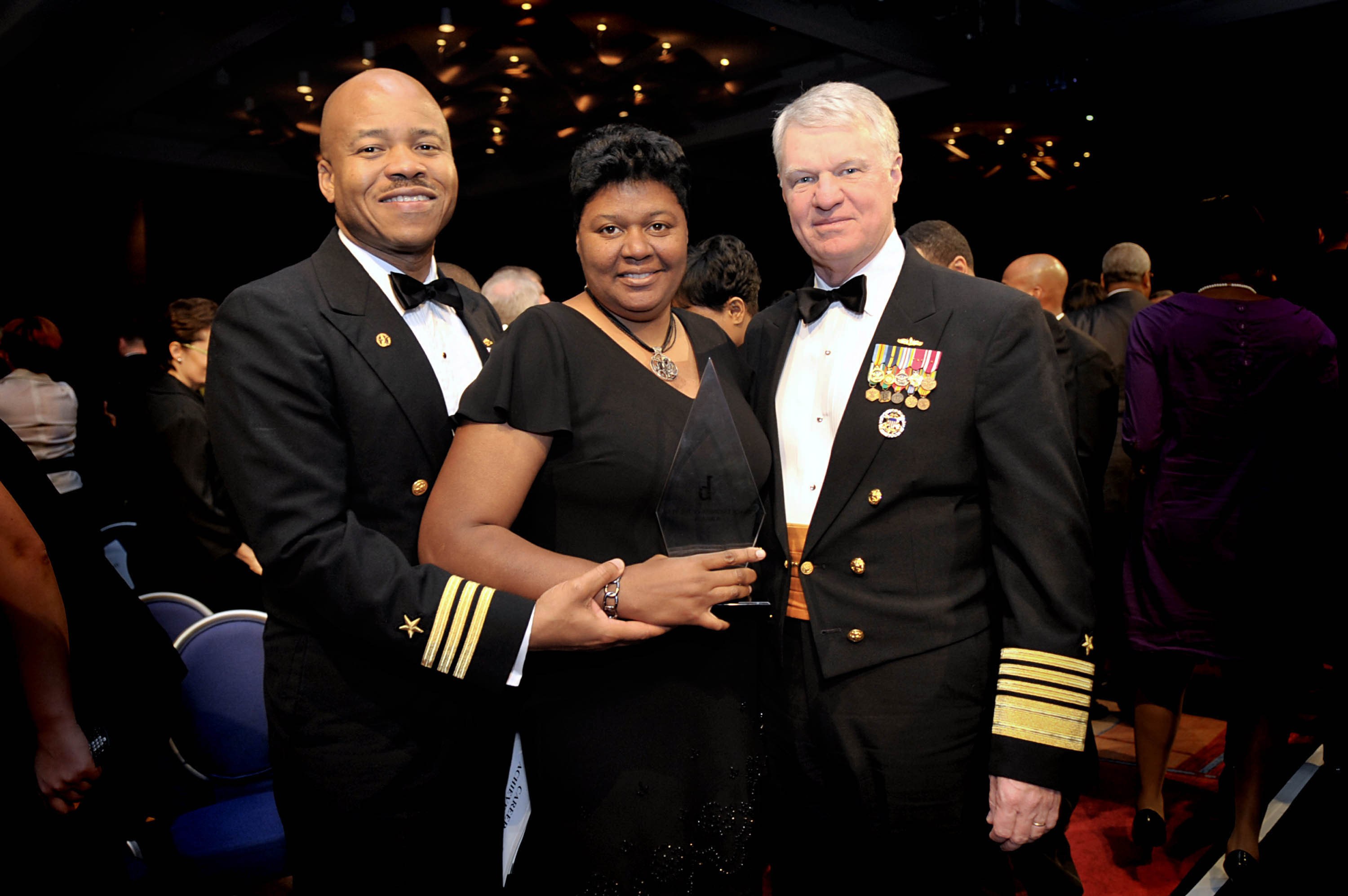 Zwei Männer in schwarzen Blazern und weißen Hemden stehen neben einer Frau in einem schwarzen Kleid, die eine Trophäe hält, auf einem roten Teppich, während andere im Hintergrund sitzen und stehen unter beleuchteter Decke.