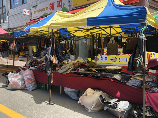 Ein Markt mit Zelten, die Taschen, Hüte und andere Artikel präsentieren, vor einem Hintergrund von Gebäuden mit Fenstern und Texttafeln.