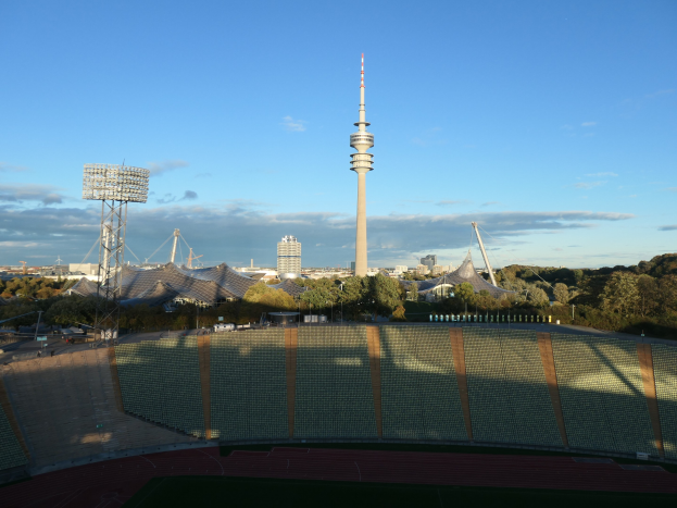 Luftaufnahme des Olympiastadions in Berlin, Deutschland, mit dem Fernsehturm (Fernsehturm) im Hintergrund, umgeben von B"umen, Geb"uden und beleuchteten Bereichen unter einem bew"olkten Himmel.