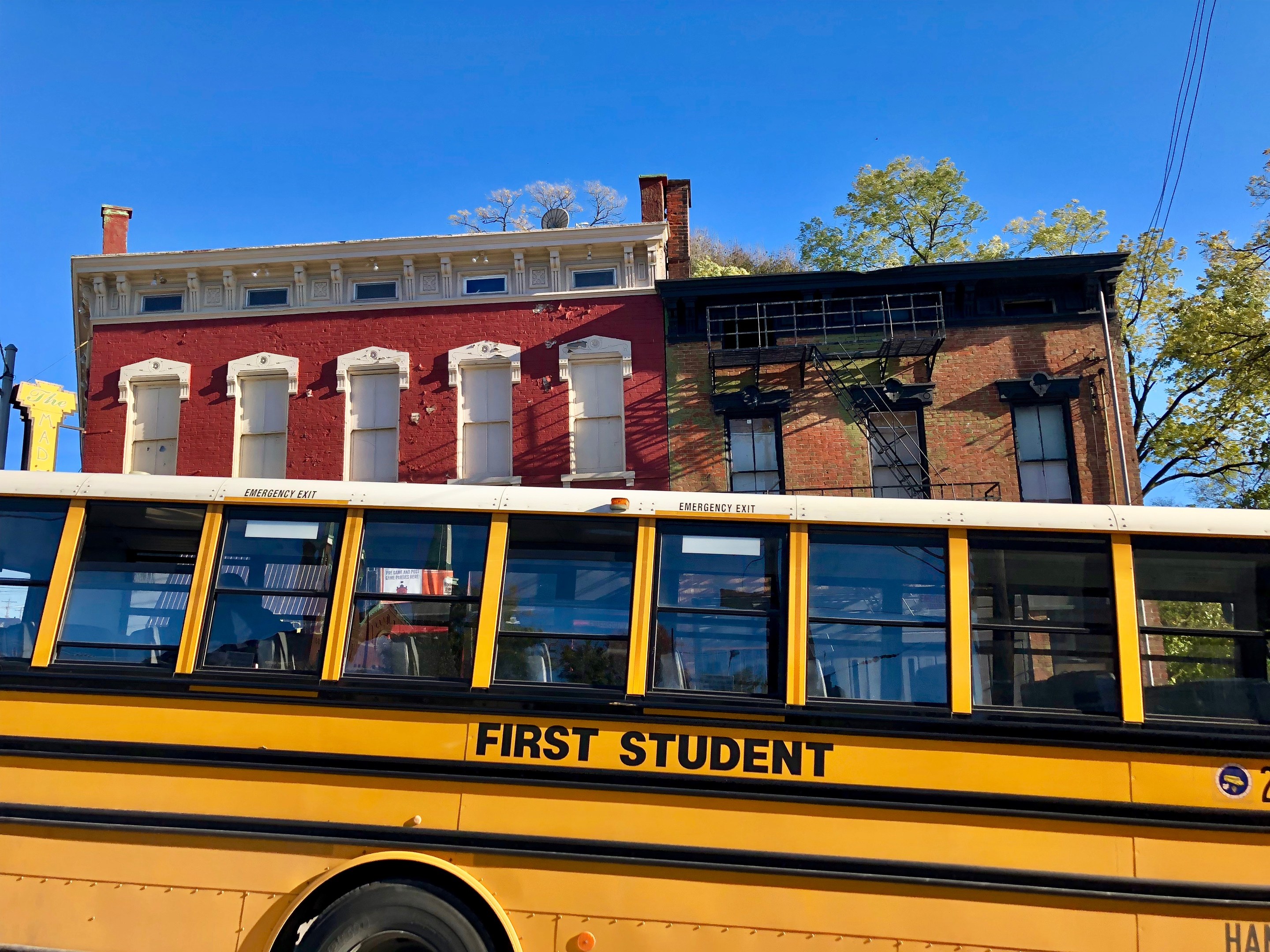 Gelber Schulbus mit der Aufschrift "First Student" vor einem roten Backsteingebäude mit ein paar Menschen drinnen, Bäumen und einem klaren blauen Himmel im Hintergrund.