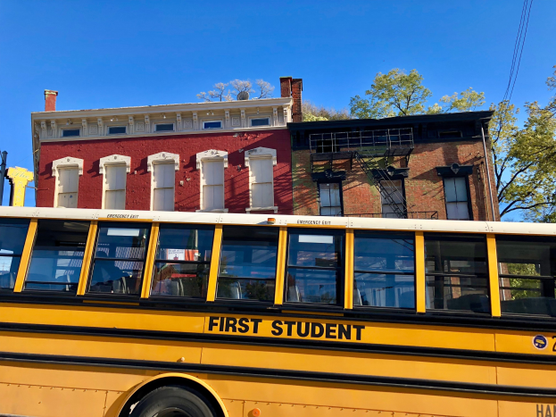 Gelber Schulbus mit der Aufschrift "First Student" vor einem roten Backsteingebäude mit ein paar Menschen drinnen, Bäumen und einem klaren blauen Himmel im Hintergrund.