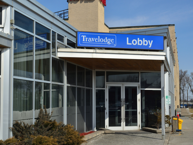 Außenansicht des Travelodge Hotels in Ottawa, Ontario, mit Glaswänden und -türen, einem Textschild, Topfpflanzen, Bäumen und einem klaren blauen Himmel.