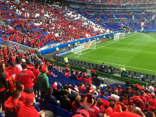 Eine große Menschenmenge in einem Stadion schaut bei einem Fußballspiel zu, mit Sitzplätzen und Stehplätzen, Fahnen, einem Torpfosten und Deckenleuchten.