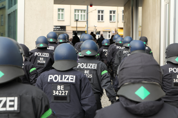 Gruppe von Polizisten in Uniform und Helmen vor einem Gebäude mit einigen Menschen und Strukturen im Hintergrund.