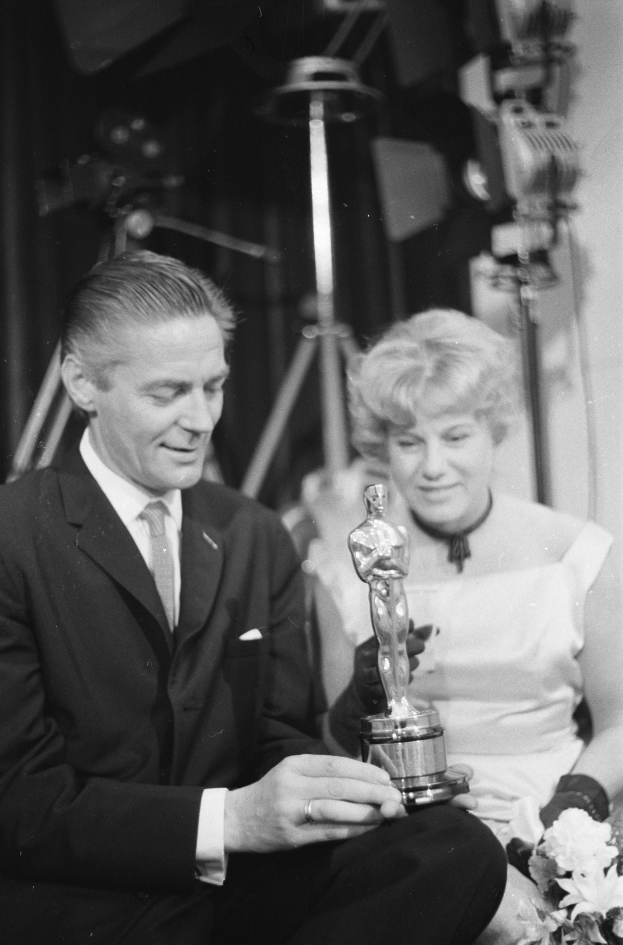 Ein Mann in einem schwarzen Blazer, weißem Hemd und Krawatte hält eine Oscar-Statue in der Hand und sitzt neben einer Frau, die einen Blumenstrauß hält, beide in formeller Kleidung, mit Lichtern im Hintergrund, in Schwarz-Weiß.