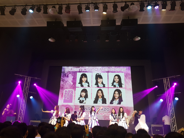 Eine Gruppe von Menschen, die auf einer hell erleuchteten Bühne performen, mit einer Menge darunter, einem Sänger mit Mikrofon und einem Musiker, der ein Instrument spielt, während ein großer Bildschirm Bilder der K-Pop-Gruppe BK48 anzeigt.